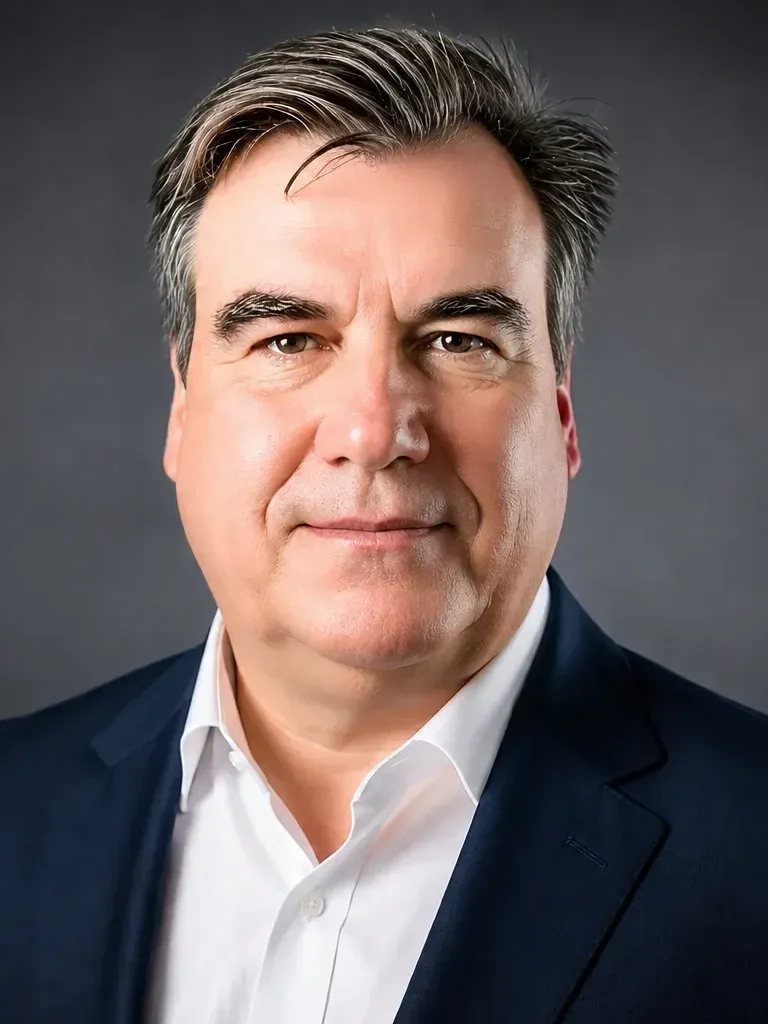 Ein Mann im dunkelblauen Anzug und weißen Hemd vor dunklem Hintergrund.